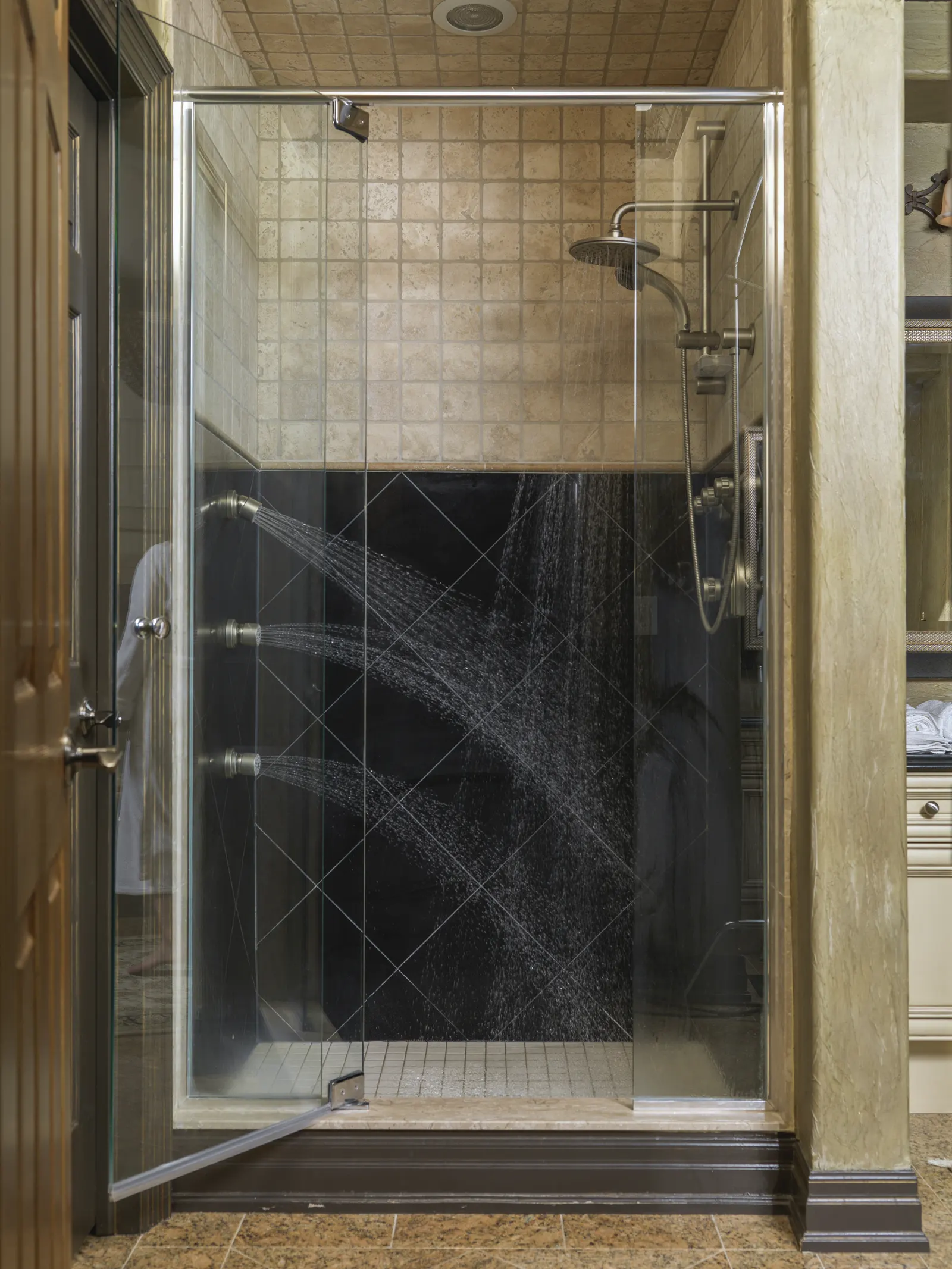 Glass shower with a hinged door ajar, running body-spray jets and travertine tile.