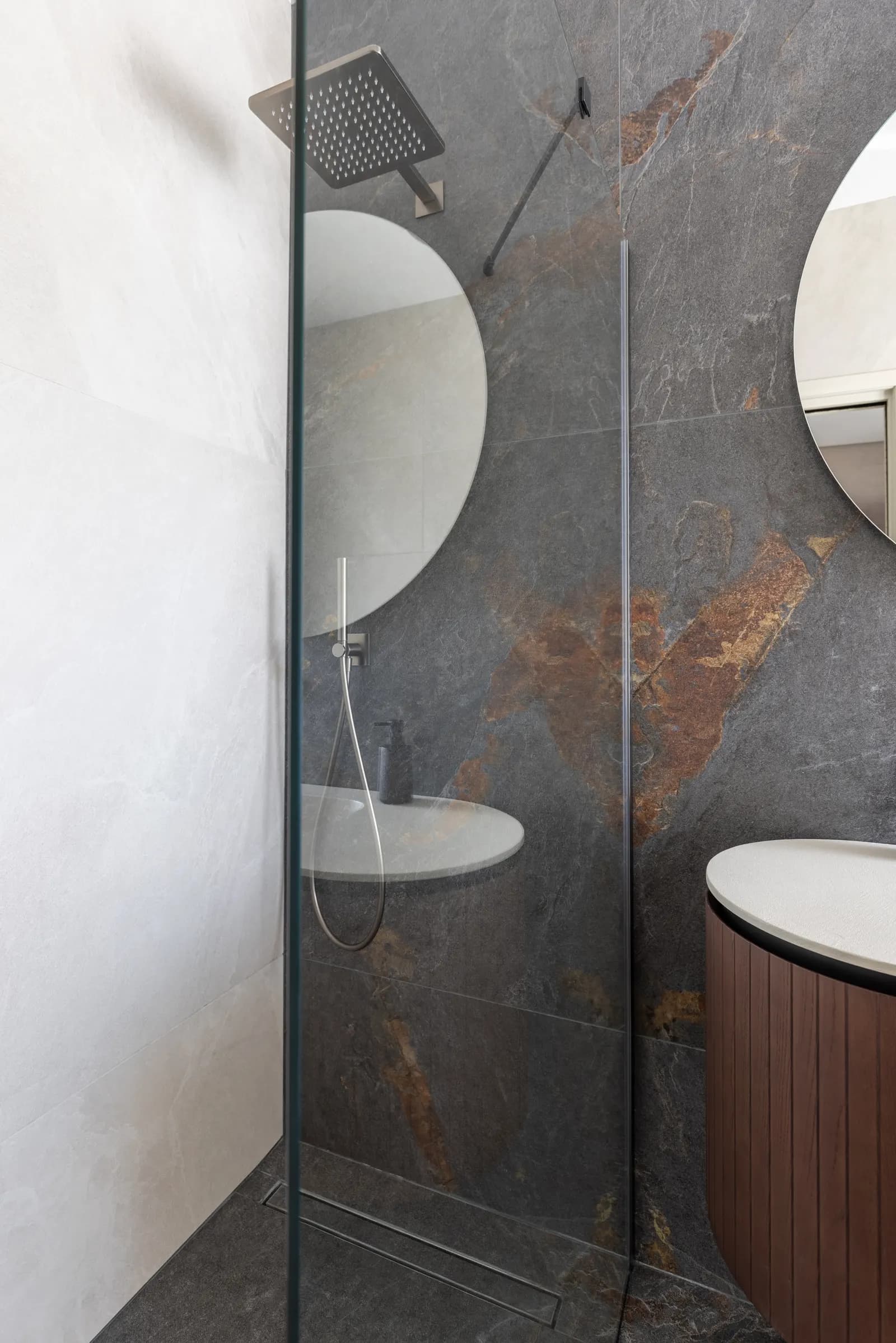 Frameless fixed glass shower screen against grey slate tile with a round mirror.