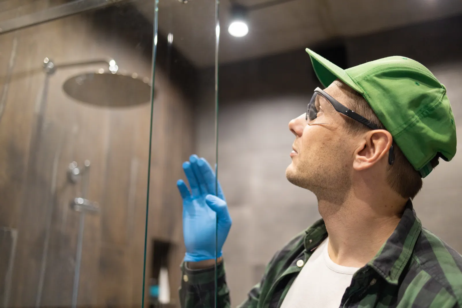 A technician in a cap and glove inspects a glass shower panel in a dark bathroom.