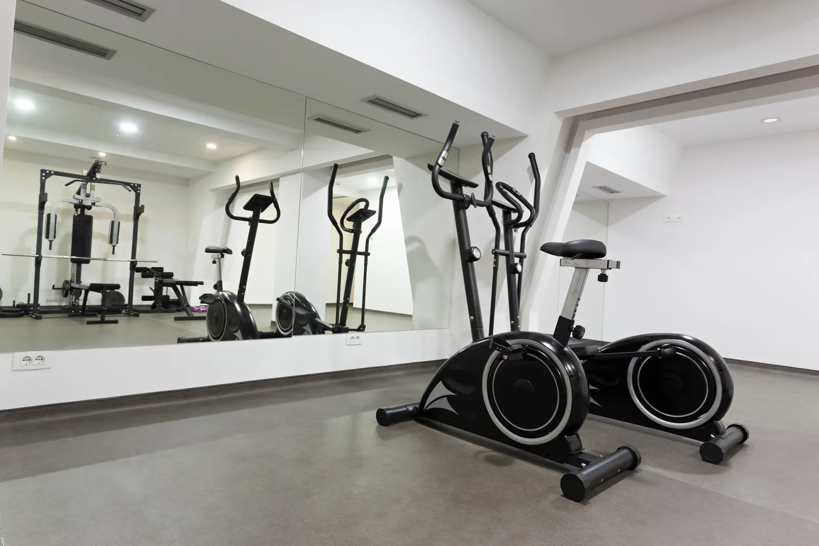 A home gym with a large wall mirror reflecting exercise bikes and weight equipment.