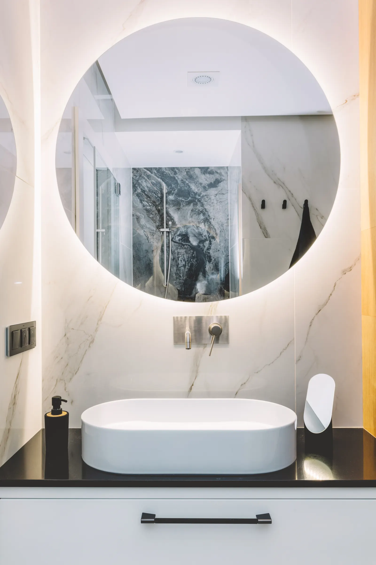 Round backlit mirror on a marble wall above a white vessel sink with a brushed metal wall faucet.