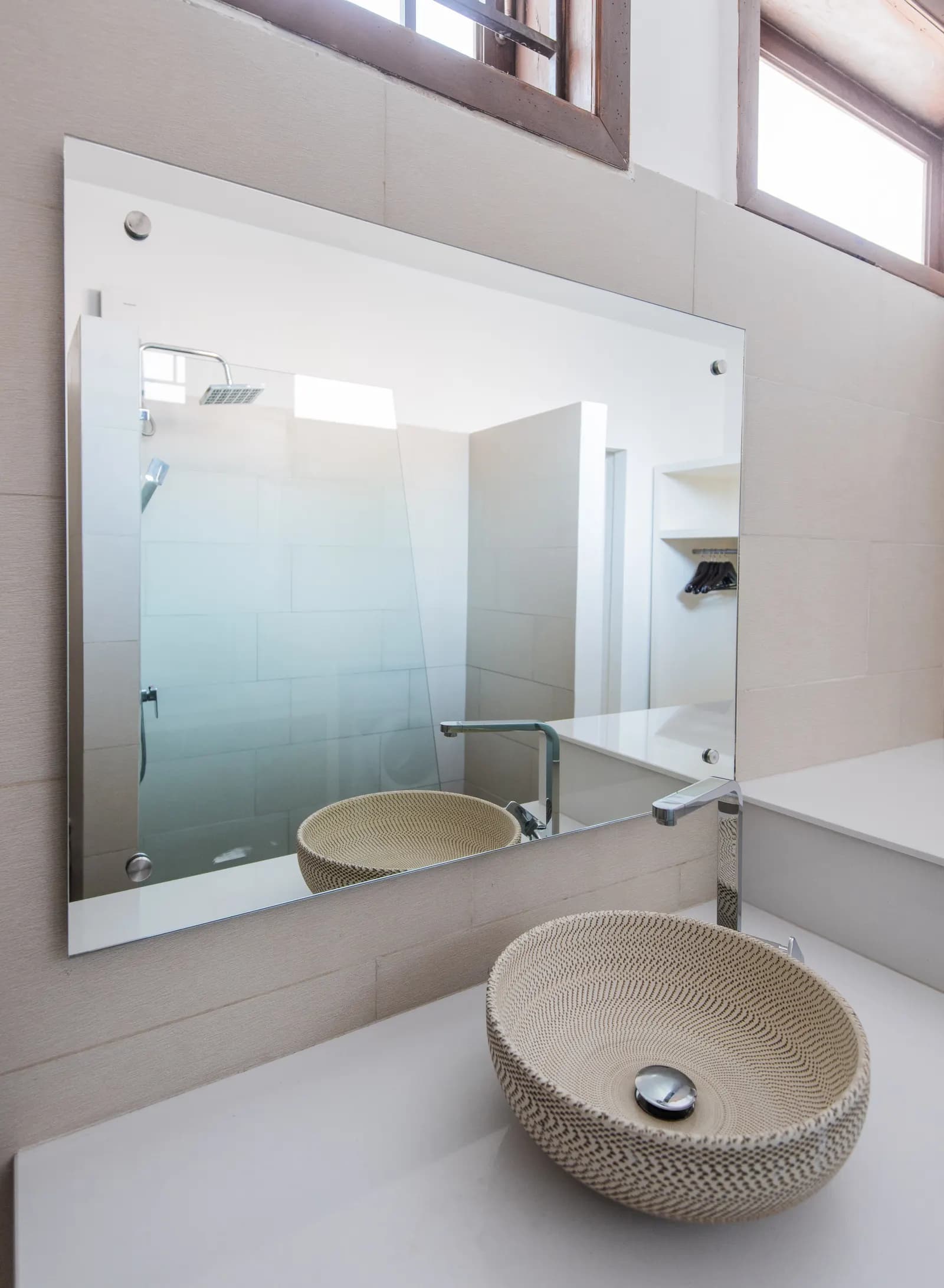 Large frameless rectangular mirror on standoff bolts above a woven bowl vessel sink.